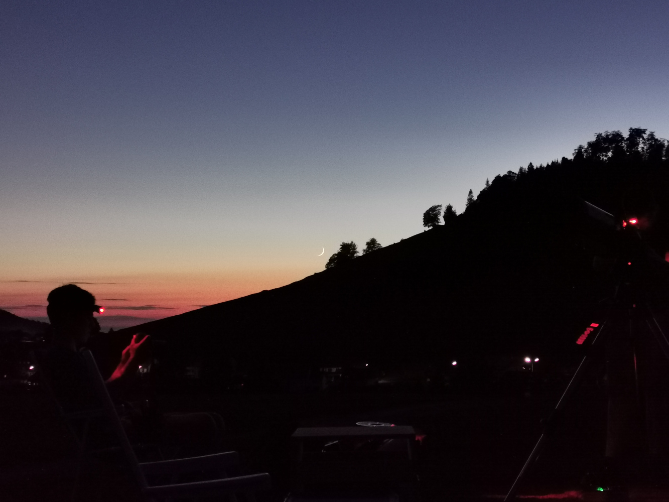 Dämmerung in Gersbach, Farben des Himmels von Dunkelblau bis Orange, Mondsichel über dunklem Horizont - Skyline wie Scherenschnitt, im Vordergrund Schüler*innen mit Teleskopen. 